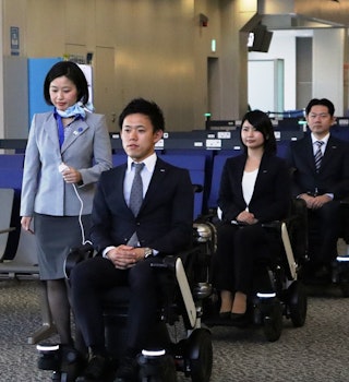 A line of three wheelchair users, accompanied by an ANA staffer, at Tokyo Narita International Airport